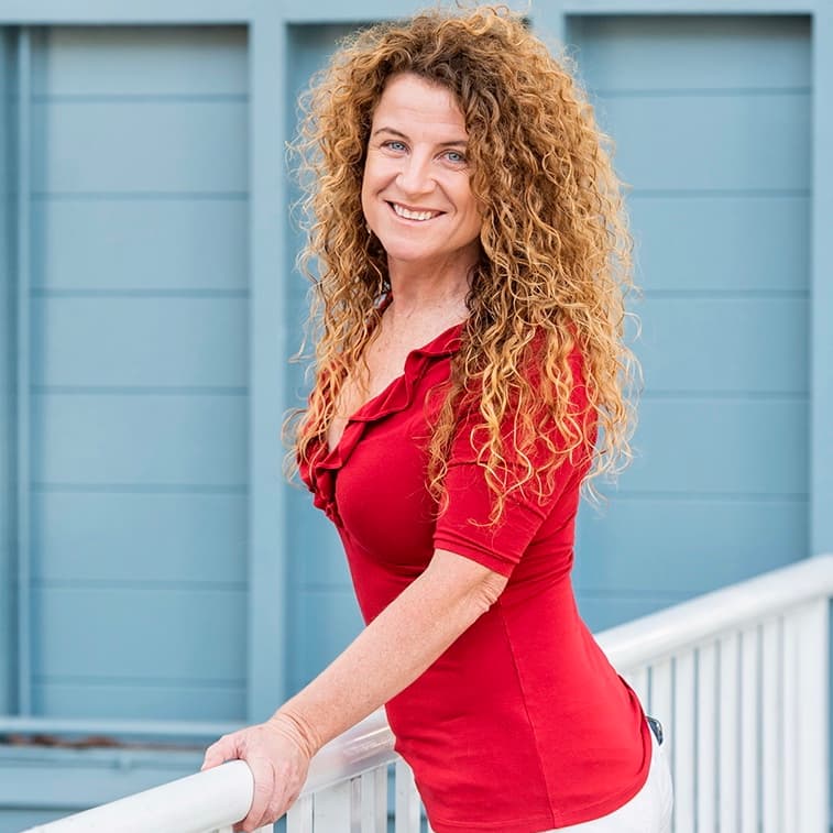 Portrait of Lisa Monks in a red top, smiling in front of a pale blue timber wall