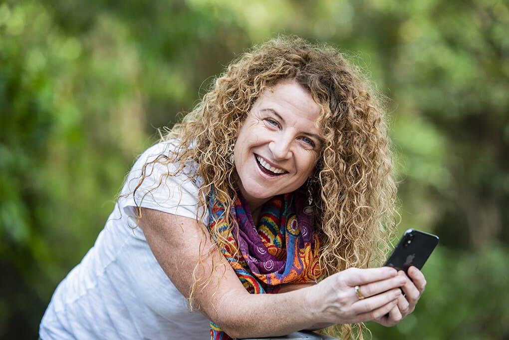 Lisa Monks smiling and holding her phone, leaning forward in a leafy outdoor setting